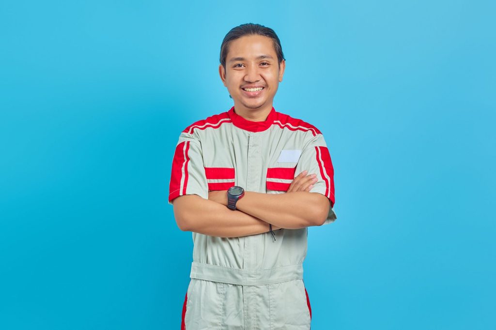 Portrait of a smiling mechanic in a studio. The man is wearing a uniform and is posed against a blue background.