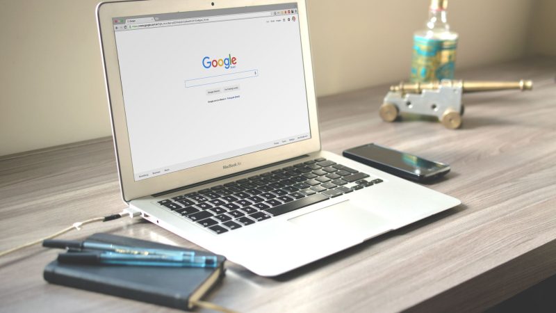 A neat workspace featuring a laptop displaying Google search, a smartphone, and a notebook on a wooden desk.